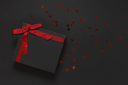 Black gift box with red ribbon and red holographic glitter confetti form of stars on dark background top view flat lay. Holiday concept birthday gift, new year or Christmas presents Xmas holiday.の写真素材