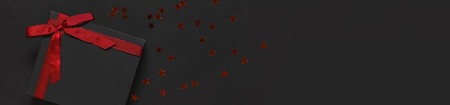 Black gift box with red ribbon and red holographic glitter confetti form of stars on dark background top view flat lay. Holiday concept birthday gift, new year or Christmas presents Xmas holiday.の写真素材