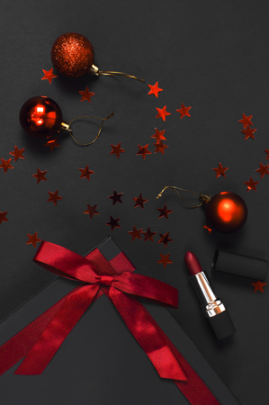 Black gift box with red ribbon, lipstick, Christmas balls, red holographic confetti on dark background top view flat lay. Holiday concept, new year presents Xmas holiday Congratulations backgroundの写真素材