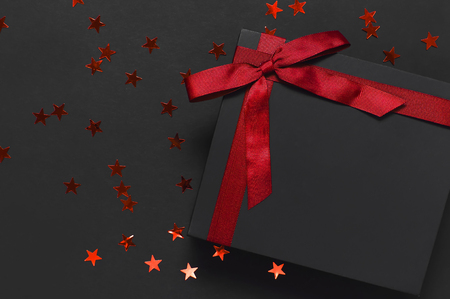 Black gift box with red ribbon and red holographic glitter confetti form of stars on dark background top view flat lay. Holiday concept birthday gift, new year or Christmas presents Xmas holiday.の写真素材