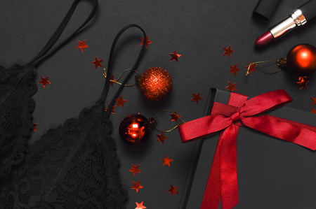 Black gift box with red ribbon, lace bra underwear, red lipstick, Christmas balls, holographic confetti on dark background top view flat lay. Female essential accessories gift for new year christmasの写真素材