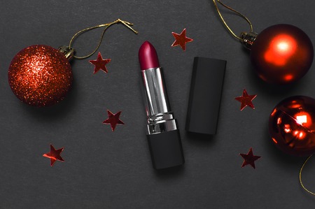 Red lipstick, Christmas balls, confetti on black background top view flat lay copy space. Professional Makeup and Beauty. Fashionable bright lipstick closeup. Make-up new year christmas conceptの写真素材