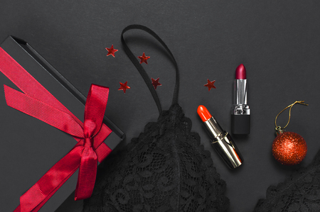 Black gift box with red ribbon, lace bra underwear, red lipstick, Christmas balls, holographic confetti on dark background top view flat lay. Female essential accessories gift for new year christmasの写真素材