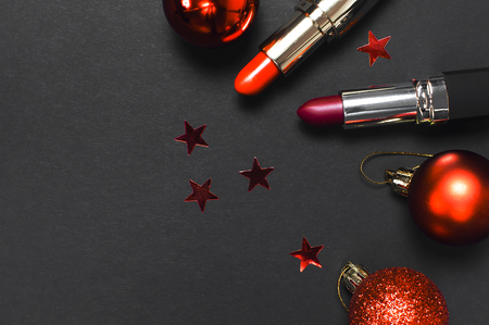 Red lipstick, Christmas balls, confetti on black background top view flat lay copy space. Professional Makeup and Beauty. Fashionable bright lipstick closeup. Make-up new year christmas conceptの写真素材