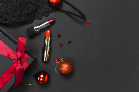 Black gift box with red ribbon, lace bra underwear, red lipstick, Christmas balls, holographic confetti on dark background top view flat lay. Female essential accessories gift for new year christmasの写真素材