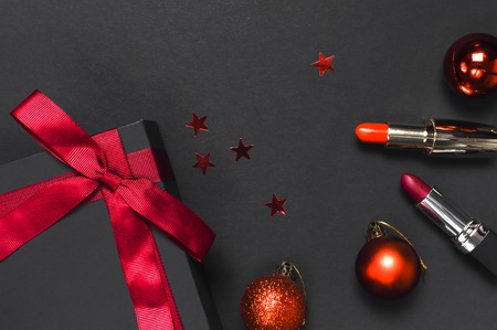 Black gift box with red ribbon, lipstick, Christmas balls, red holographic confetti on dark background top view flat lay. Holiday concept, new year presents Xmas holiday Congratulations backgroundの写真素材