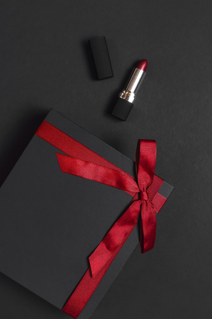 Black gift box with red ribbon, lipstick on dark background top view flat lay. Holiday concept, birthday gift, Valentine's Day, presents. Congratulations background. Beautiful dark Make-up conceptの写真素材