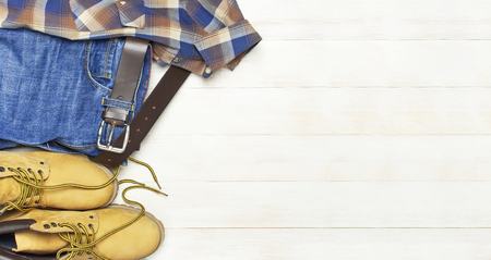 Men's casual wear, yellow work boots from natural nubuck leather, blue jeans, checkered shirt and brown belt on wooden white background top view flat lay copy space. Trendy casual shoes, youth styleの写真素材