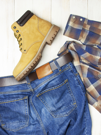 Men's casual wear, yellow work boots from natural nubuck leather, blue jeans, checkered shirt and brown belt on wooden white background top view flat lay copy space. Trendy casual shoes, youth styleの写真素材