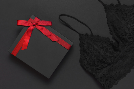 Black gift box with red ribbon, lace bra underwear, red lipstick, holographic confetti on dark background top view flat lay. Female essential erotic accessories, fashionable underwear gift to womanの写真素材