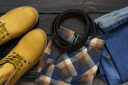 Men's casual wear, yellow work boots from natural nubuck leather, blue jeans, checkered shirt and brown belt on dark wooden background top view flat lay copy space. Trendy casual shoes, youth styleの写真素材