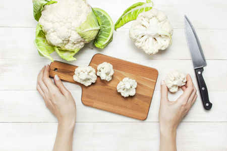 Female hands cut cauliflower with knife on cutting board, kitchen towel on rustic white wooden background top view flat lay copy space. Cooking, healthy wholesome food, vegetable, diet conceptの写真素材