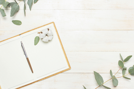 Open notebook with blank pages, pen, eucalyptus twig and cotton flowers on white wooden background top view flat lay. Fashion female blogger working desk. Cotton flowers Lifestyle gentle backgroundの写真素材