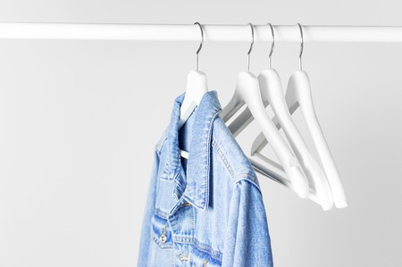 Blue denim jacket on white wooden coat hanger on a rod against light gray wall flat lay copy space. Denim, fashionable jacket, womens or mens trend clothing, fashion background. Store concept, saleの写真素材