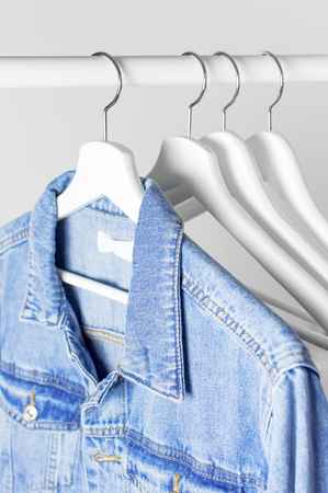 Blue denim jacket on white wooden coat hanger on a rod against light gray wall flat lay copy space. Denim, fashionable jacket, womens or mens trend clothing, fashion background. Store concept, saleの写真素材