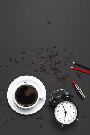 Flat lay cup black coffee, coffee beans, black alarm clock, red lipstick, color pencils on gray dark background top view copy space. Concept time to work female desktop Minimalistic coffee backgroundの写真素材