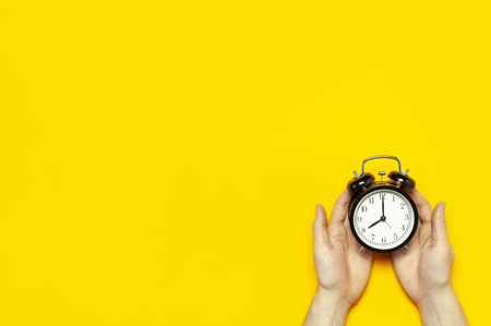 Flat lay top view male hand holding black vintage alarm clock on bright yellow color background with copy space. Creative Time concept minimal style, morning time to work.の写真素材