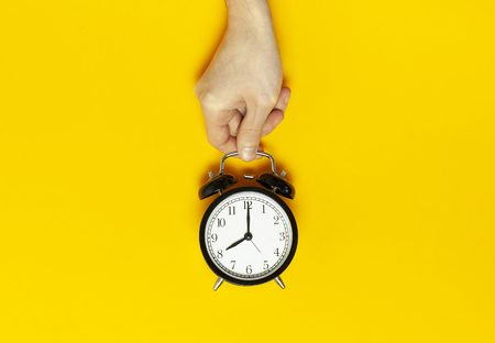 Flat lay top view male hand holding black vintage alarm clock on bright yellow color background with copy space. Creative Time concept minimal style, morning time to work.の写真素材