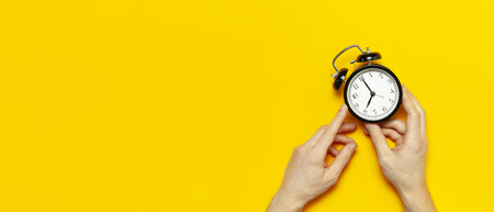 Flat lay top view male hand holding black vintage alarm clock on bright yellow color background with copy space. Creative Time concept minimal style, morning time to work.の写真素材