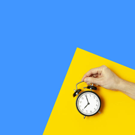 Flat lay top view male hand holding black vintage alarm clock on bright yellow blue color background with copy space. Creative Time concept minimal style, morning time to work.の写真素材