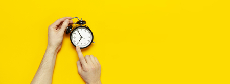 Flat lay top view male hand holding black vintage alarm clock on bright yellow color background with copy space. Creative Time concept minimal style, morning time to work.の写真素材