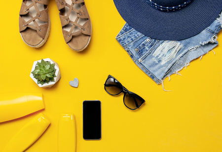 Female summer fashion background. Clothes and accessories on yellow background. Blue hat, denim shorts, sandals, sunglasses, sunscreen, phone. Flat lay top view, beauty or fashion blog concept.の写真素材