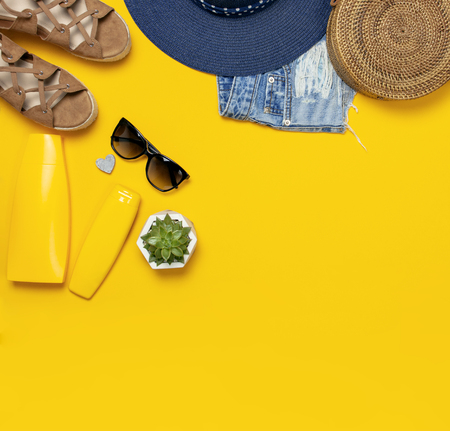 Female summer fashion background. Clothes and accessories on yellow background. Blue hat, denim shorts, round rattan bag, sandals, sunglasses, sunscreen. Flat lay top view beauty fashion blog concept.の写真素材