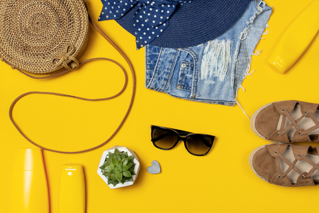 Female summer fashion background. Clothes and accessories on yellow background. Blue hat, denim shorts, round rattan bag, sandals, sunglasses, sunscreen. Flat lay top view beauty fashion blog concept.の写真素材