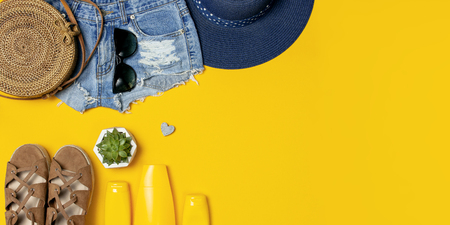 Female summer fashion background. Clothes and accessories on yellow background. Blue hat, denim shorts, round rattan bag, sandals, sunglasses, sunscreen. Flat lay top view beauty fashion blog concept.の写真素材