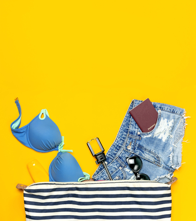 Female summer clothing accessories on yellow background. Concept of travel vacation sea Striped Nautical Bag swimming suit denim shorts sunglasses sunscreen passport phone self-stick Flat lay top view.の写真素材