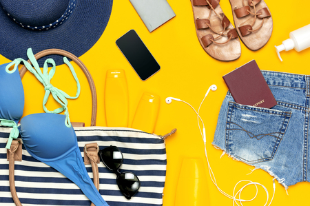 Female summer clothing accessories on yellow background. Concept of travel vacation sea. Hat swimming suit denim shorts leather sandals sunglasses sunscreen sea bag passport phone. Flat lay top view.の写真素材