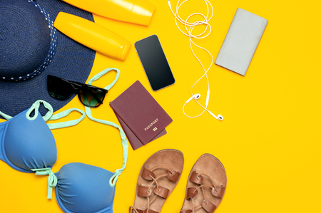 Female summer clothing accessories on yellow background. Concept of travel vacation sea. Blue hat swimming suit leather sandals sunglasses sunscreen passport phone headphones Flat lay top view.の写真素材