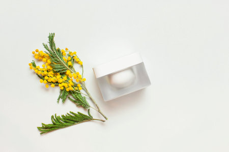 White cosmetic bottle containers with yellow Mimosa flowers on light background top view flat lay....