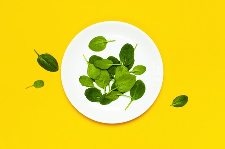 Fresh green spinach leaves on white plate on bright yellow background flat lay top view copy space. Baby young spinach leaves, Ingredient for salad, healthy food, diet. Creative food conceptの写真素材