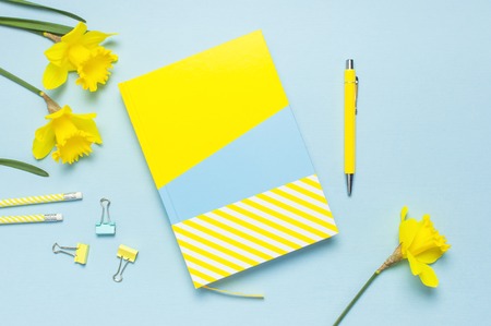 Yellow-blue notebook, pen, clips, spring flowers daffodils narcissus on blue background. Female desktop, Office desk, spring concept. Flat lay, top view, copy space. Template for feminine blog.の写真素材