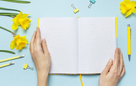 Women's hands, open blank notebook, pen, clips, spring flowers daffodils narcissus on blue background. Female desktop, Office desk, spring concept. Flat lay, top view, copy space. Template for blog.の写真素材
