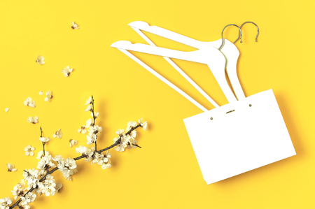 White wooden hangers, spring flowering branch, white gift bag on yellow background. Spring sale concept discount store shopping empty hanger. Creative fashion banner. Flat lay top view copy spaceの写真素材