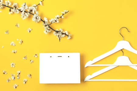White wooden hangers, spring flowering branch, white gift bag on yellow background. Spring sale concept discount store shopping empty hanger. Creative fashion banner. Flat lay top view copy spaceの写真素材