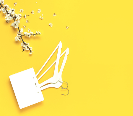 White wooden hangers, spring flowering branch, white gift bag on yellow background. Spring sale concept discount store shopping empty hanger. Creative fashion banner. Flat lay top view copy spaceの写真素材