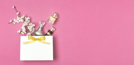 White gift bag with gold ribbon, bottle of perfume, spring white flowers on bright pink background. Greeting card with delicate flowers Pink floral background. Spring minimal concept Flat lay top view.の写真素材