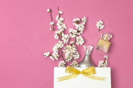 White gift bag with gold ribbon, bottle of perfume, spring white flowers on bright pink background. Greeting card with delicate flowers Pink floral background. Spring minimal concept Flat lay top view.の写真素材