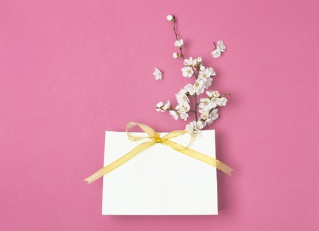White gift bag with gold ribbon and branch of spring white flowers on bright pink background. Greeting card with delicate flowers Pink floral background. Spring minimal concept. Flat lay top view.の写真素材