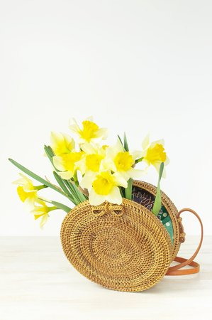 Fashionable handmade natural organic round rattan bag, yellow narcissus or daffodil flowers on light wooden background flat lay. Trendy bamboo bag Ecobags from Bali. Summer spring fashion concept.の写真素材