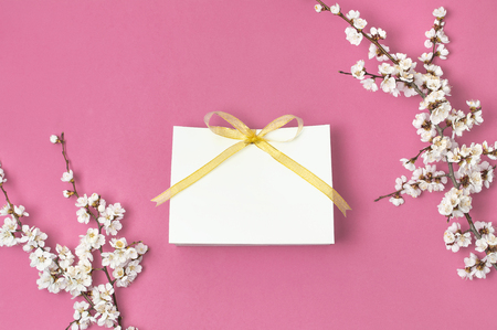 White gift bag with gold ribbon and branch of spring white flowers on bright pink background. Greeting card with delicate flowers Pink floral background. Spring minimal concept. Flat lay top view.の写真素材