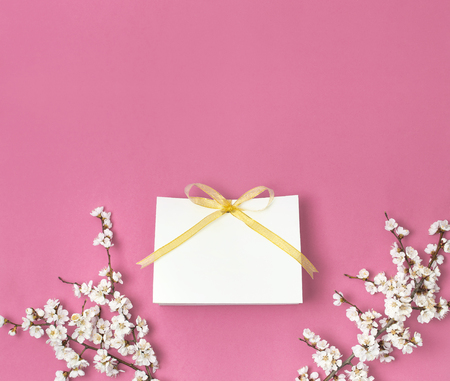 White gift bag with gold ribbon and branch of spring white flowers on bright pink background. Greeting card with delicate flowers Pink floral background. Spring minimal concept. Flat lay top view.の写真素材