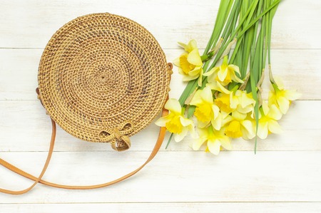 Fashionable handmade natural organic round rattan bag, yellow narcissus or daffodil flowers on light wooden background flat lay. Trendy bamboo bag Ecobags from Bali. Summer fashion concept Top view.の写真素材