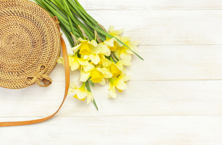 Fashionable handmade natural organic round rattan bag, yellow narcissus or daffodil flowers on light wooden background flat lay. Trendy bamboo bag Ecobags from Bali. Summer fashion concept Top view.の写真素材