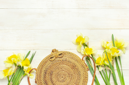 Fashionable handmade natural organic round rattan bag, yellow narcissus or daffodil flowers on light wooden background flat lay. Trendy bamboo bag Ecobags from Bali. Summer fashion concept Top view.の写真素材