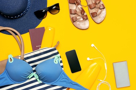 Female summer clothing accessories on yellow background. Concept of travel vacation sea. Hat swimming suit denim shorts leather sandals sunglasses sunscreen sea bag passport phone. Flat lay top view.の写真素材