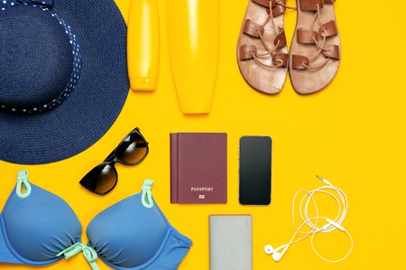 Female summer beach accessories on yellow background. Concept of travel vacation sea. Blue hat swimming suit leather sandals sunglasses sunscreen passport phone headphones Flat lay top view.の写真素材